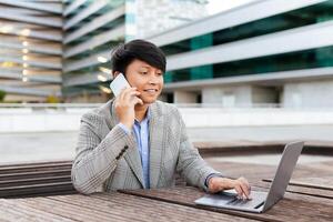 A young Asian man in a stylish suit engages in a phone conversation while using a laptop at an outdoor workspace. The modern background features buildings and a sleek environment. photo