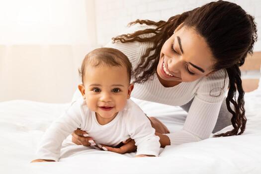 africano madre jugando con linda bebé hija Ayudar su gatear en cama en dormitorio a hogar. joven mamá disfrutando maternidad rutina y niño cuidado. maternidad salir estilo de vida foto