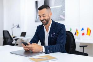 A well-dressed man smiles while using his smartphone at a sleek office desk, decorated with national flags. The bright workspace evokes a professional and inviting atmosphere. photo