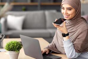 Work From Home. Young Muslim Business Lady Using Voice Search Assistant, Talking On Mobile Phone And Using Laptop Computer. Smiling Woman Sitting At Desk With Pc At Home Office, Wearing Hijab photo