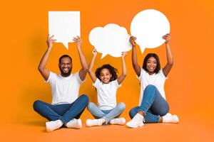 Diverse Thoughts. Smiling black family of three holding empty speech bubbles with different shape above their heads, sitting on floor over yellow studio background. Free place for text, copy space photo
