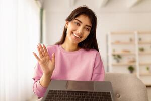 Greeting Concept. Portrait of smiling beautiful woman waving hello with hand looking at camera, using laptop at home office. Cheerful lady saying hi or goodbye during call, blurred background photo