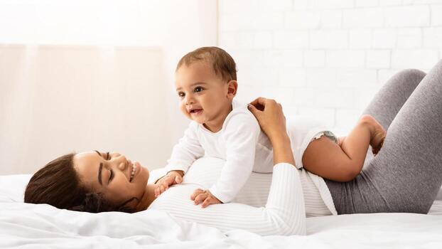 contento negro mamá jugando con bebé chico acostado en su estómago en dormitorio a hogar. africano americano mamá disfrutando maternidad gasto hora con niñito infantil durante maternidad salir. lado ver foto