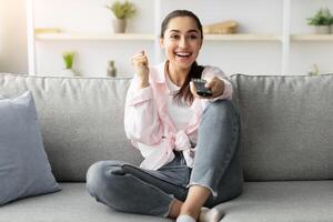 Having Fun. Portrait of optimistic enthusiastic Excited young woman watching TV show or movie, getting emotional over plot twist, shaking clenched fists, making winner gesture, sitting on sofa photo