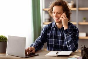 Cheerful Millennial Businessman Talking On Cellphone Working Online On Laptop Sitting At Workplace In Modern Office. Freelancer Man Chatting On Phone Indoors. Mobile Communication. Side View photo