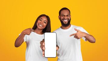 Advertising New Mobile App. Portrait of smiling black couple holding cellphone with empty screen in hands, showing and pointing fingers at device with blank space for mockup, yellow studio background photo