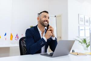 A smiling businessman is speaking on the phone at his desk, surrounded by flags and modern decor. He appears focused while managing his work tasks on a laptop. photo