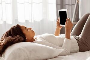 Free Time At Home And Weekend Pastime Concept. Young Black Lady Holding And Using Digital Tablet Computer With Blank Monitor For Mock Up, Reading News Or Electronic Book, Lying In Bed, Free Space photo