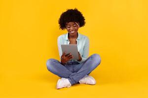 Smiling African American Female Using Tablet Computer Browsing Internet Sitting On Floor Over Pink Background In Studio. E-Learning, Networking And Online Technology. Gadget Lifestyle Concept photo