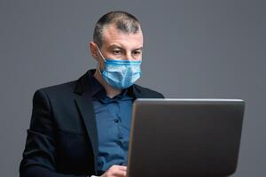 Entrepreneur Man Working Using Laptop Computer Wearing Protective Mask Sitting On Gray Background. Distant Work Concept. Studio Shot photo