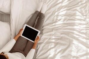 Above Top High Angle View Of Young Black Woman Using Digital Tablet With Empty Screen Reading E-Book Online, Sitting On Bed At Home On Weekend. Internet Website Advertising, Mock Up, Free Copy Space photo