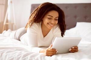 Portrait of happy African American woman lying in bed and smiling, holding and using digital tablet, chatting or browsing social media. Cheerful lady reading news or internet blog, watching photo