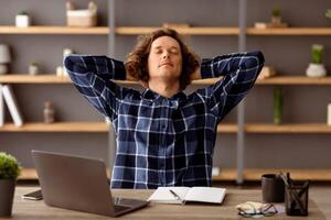 Successful Freelancer Guy At Laptop Resting Holding Hands Behind Head Relaxing At Workplace After Online Work Sitting At Desk In Modern Office. Selective Focus photo