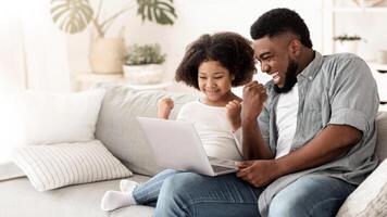 Excited black father and little daughter playing computer games on laptop at home, celebrating win with clenched fists, sitting on couch, copy space photo