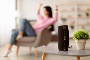 Modern Technology Concept. Closeup of smart portable wireless speaker on the table, selective focus. Young woman listening to music and dancing, sitting on armchair in the blurred background photo