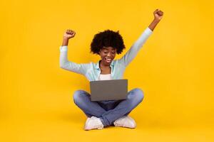 Excited African Woman With Laptop Shaking Fists In Joy And Gesturing Yes Celebrating Great News Sitting On Floor In Studio Over Pink Background. Joyful Lady Posing With Computer. Internet And Gadgets Concept photo