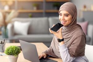 Digital Speaker Application. Portrait of young muslim woman in hijab sitting at table, talking on mobile phone, using laptop and virtual voice assistant for online search or to record reminder photo