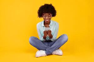 Cheerful Black Female Using Smartphone Texting And Networking In Social Media Sitting Over Pink Background In Studio. Bushy African American Young Woman Using Mobile Application On New Cellphone photo
