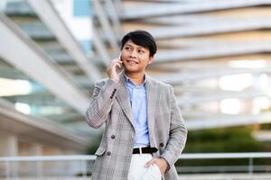 A young Asian man dressed in a fashionable blazer enjoys a phone conversation while standing outside a contemporary building in an urban setting, exuding confidence and style. photo