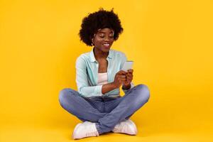 Happy African Woman Using Phone Texting And Browsing Internet Sitting On Floor Over Pink Background. Bushy Black Lady Networking In Social Media Using Smartphone App. Mobile Application For Cellphone photo