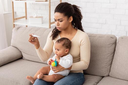 enfermo bebé teniendo fiebre. preocupado negro mamá medición pequeño del niño temperatura con termómetro cuidando para enfermo hijo sentado en sofá a hogar. niño cuidado y infantil salud problema concepto foto