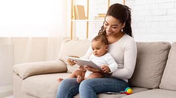 Cheerful Black Mommy Using Digital Tablet Holding Little Baby Showing Cartoons Online Sitting On Sofa In Living Room At Home. Mother Browsing Internet On Computer Using Games Application For Infant photo