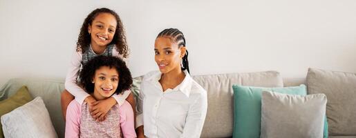 A black mother sits on a sofa with her two daughters, smiling joyfully. The daughters are playfully positioned, one on the mother lap and the other behind her, creating a warm family moment. photo