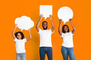 Happy black family of three in white t-shirts holding empty speech bubbles with different shape above their heads, standing isolated at yellow studio. Free place for text, copy space. Diverse Thoughts photo