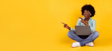 Cheerful Black Woman With Laptop Pointing Fingers Aside At Empty Space For Advertisement Text Posing Sitting On Floor Over Pink Background. Studio Shot. Look There Concept photo