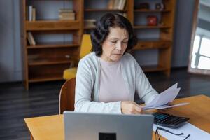 Middle aged senior woman sit with laptop and paper document. Pensive older mature lady reading paper bill pay online at home managing bank finances calculating taxes planning loan debt pension payment photo