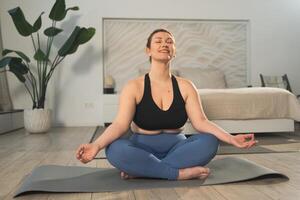 Yoga mindfulness meditation. Woman practicing yoga in living room at home. Plus size female sitting in lotus pose meditating smiling relaxing indoor. Girl doing breathing practice. Yoga at home photo
