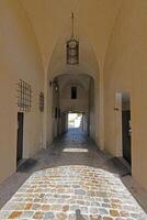 An arched stone corridor with a cobblestone floor, dim lighting, and metal lanterns hanging from the ceiling photo