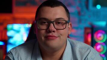 A man with glasses and a shirt in front of a neon light video