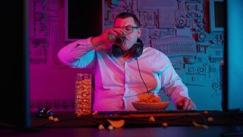 A man in glasses drinking from a glass of beer while sitting at a table with a computer video