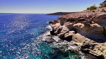 Sunlight glistens on a stunning turquoise sea as gentle waves lap against a rugged coastline under a clear summer sky video