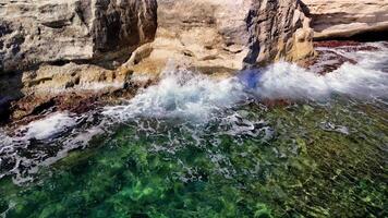 Turquoise sea waves crashing against rugged rocks on a sunny day create a stunning display of white foam, highlighting the vibrant, clear water and showcasing nature's raw energy video