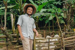 Cheerful Asian Muslim man standing in front of livestock goats for sacrifices behind. Eid Al Adha concept. photo