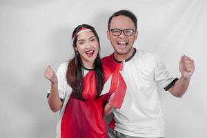 Cheerful Indonesian young couple holding flags while raising fist to support in a sport match. photo
