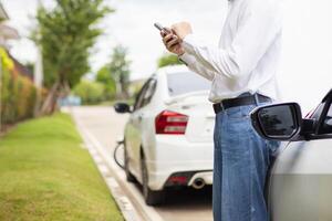 young man is standing in front of the hood of a car with engine problems and using his mobile phone to search for a local car service center and notify the mechanic to come and check the car. photo