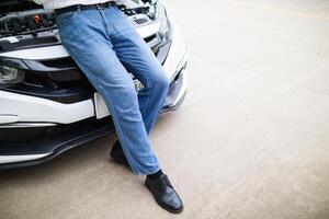 young man is standing in front of the hood of a car with engine problems and using his mobile phone to search for a local car service center and notify the mechanic to come and check the car. photo