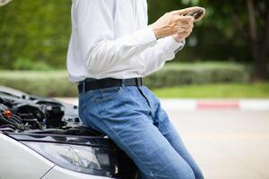 young man is standing in front of the hood of a car with engine problems and using his mobile phone to search for a local car service center and notify the mechanic to come and check the car. photo