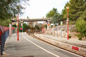 Spain, Valencia, March 29, 2024. Train Platform at a Quiet Station Surrounded by Greenery, Under a Bridge, on a Sunny Day photo