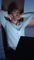 A man relaxes by the ocean at dusk, working on his laptop and enjoying the beautiful view video