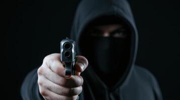 masked individual in black hoodie points gun directly at camera, creating tense and threatening atmosphere. dark background enhances sense of danger and mystery photo