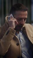 A man dressed in a beige coat is talking on his phone while standing in an urban setting video