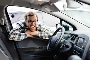 Young man looking through window inside new car, checking it before purchase at modern dealership store. Cheerful guy buying or renting automobile at showroom photo