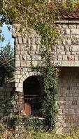 Rustic stone building covered in creeping ivy with an arched window and metal railing. The structure appears old and weathered. photo