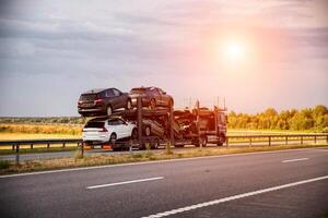 An auto transporter semi-trailer truck transports broken damaged cars from Europe in the summer against a sunset background. Car import and export concept, shipping photo