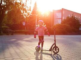 A caucasian girl of seven years in a pink protective outfit jumps, laughs and fools around near a scooter in the summer against the backdrop of a sunset. photo