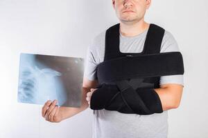 A man in a black bandage supporting the shoulder joint after surgery holds in his hand an x-ray of a dislocated shoulder and a broken arm. Rehabilitation after a clavicle fracture injury. photo